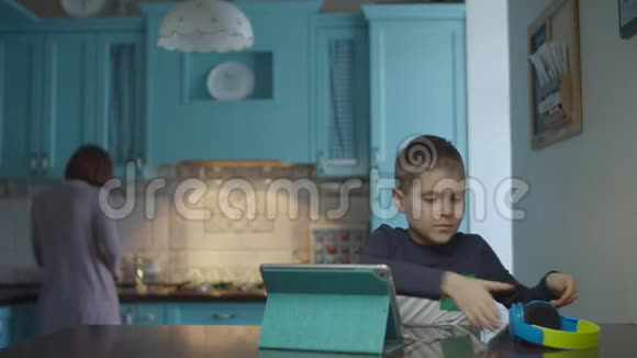 自闭症男孩在厨房里使用带耳机的平板电脑而母亲则带零食患有自闭症的孩子视频的预览图