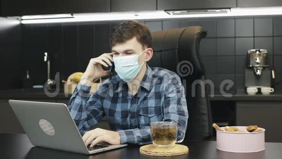 戴防护面罩的人在家打电话男性自由职业者带医疗面具在家工作手提电脑视频的预览图