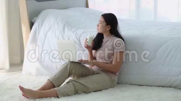 美丽的亚洲年轻女性坐在床边在卧室里喝牛奶看着外面视频的预览图