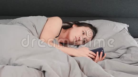 年轻女孩躺在灰色的床上在智能手机上打字的肖像布鲁内特女士躺在空床上看着智能手机魅力视频的预览图