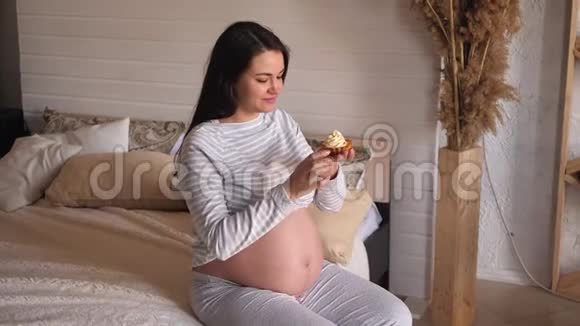 美丽的年轻孕妇在家里的床上吃蛋糕微笑着视频的预览图