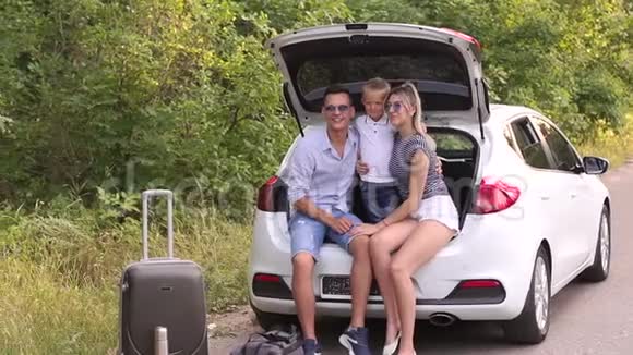 父母亲吻他们心爱的儿子坐在路边的汽车后备箱里视频的预览图