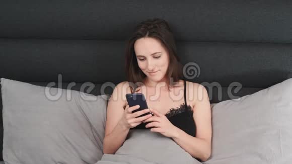 微笑的女人看着躺在床上的智能手机屏幕年轻迷人的黑发女性躺在灰色的床上手里拿着电话视频的预览图