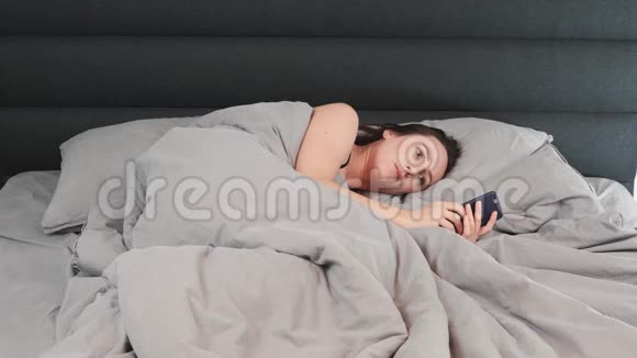 布鲁内特女士躺在床上看着智能手机屏幕年轻孤独的女性独自躺在床上手里拿着电话视频的预览图