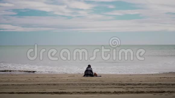 在风和日丽的天气里坐在海边的女人裹着舒适温暖的格子呢大海上有波浪后景总计划视频的预览图
