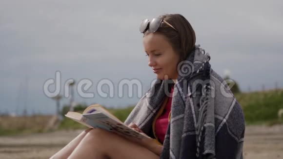 一位年轻的浪漫女子在海边看书时被一个格子包裹的肖像视频的预览图