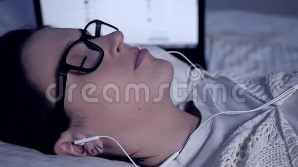 女孩晚上听音乐戴着眼镜和耳机的迷人年轻女子躺在笔记本电脑旁边的床上她喜欢视频的预览图