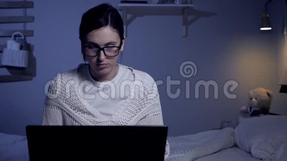 女孩晚上在电脑上工作年轻漂亮的戴眼镜的女人学生或自由职业者坐在黑暗的床上视频的预览图