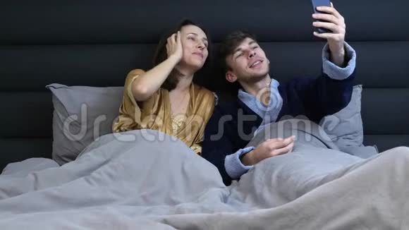 现代家庭在智能手机上自拍躺在灰色的床上年轻的已婚夫妇在酒店的床上自拍有趣的女性和男性视频的预览图