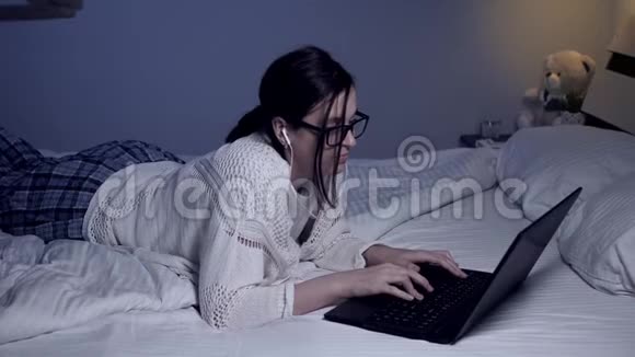戴着耳机眼镜的女孩独自躺在公寓的床上听音乐在笔记本电脑上打字学生视频的预览图