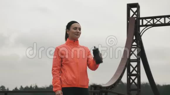 女运动员在公园户外喝水年轻的运动女郎穿着鲜艳的运动服从瓶子里喝能量饮料视频的预览图