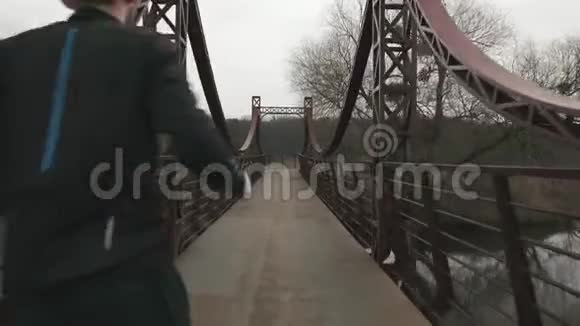 瘦小的男人在公园的桥上横渡河背面的景色男运动员在户外跑步穿黑色紧身衣的年轻人视频的预览图