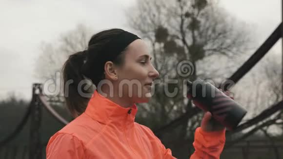 年轻女性从瓶子里喝能量饮料的肖像布鲁内特女运动员在公园里从室外的瓶子里喝水女孩视频的预览图