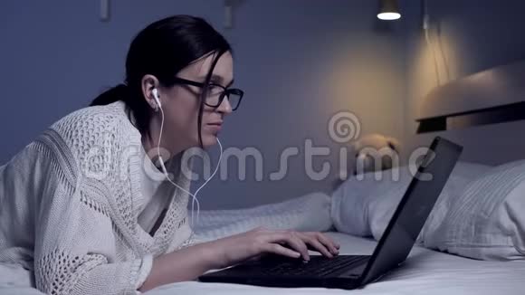 戴着耳机眼镜的女孩躺在床上听音乐在笔记本电脑上打字学生自由职业者工作视频的预览图