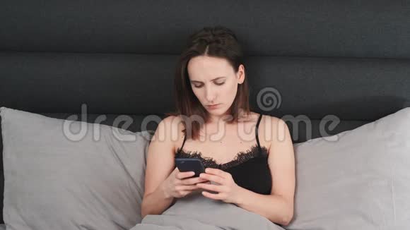 悲伤的黑发女人正在家里的床上看智能手机屏幕年轻的女性躺在灰色的床上打电话视频的预览图