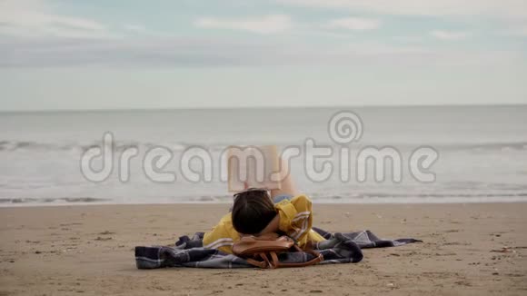 穿着黄色夹克和牛仔裤短裤的布鲁内特女士在海边看书放松大海上有波浪后视图视频的预览图