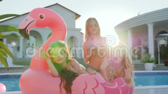 可爱的女孩躺在游泳池附近的充气粉红火烈鸟上被宠坏的富有的孩子在户外背光孩子们玩得开心视频的预览图