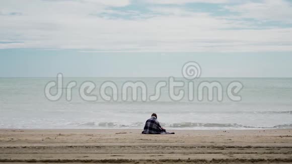 在风和日丽的天气里坐在海边的女人裹着舒适温暖的格子呢大海上有波浪后景总计划视频的预览图