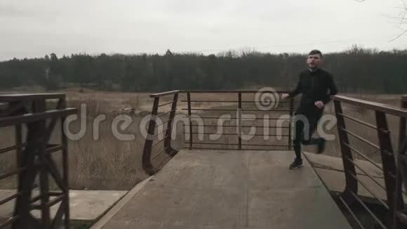 男子在桥上横渡河前景白人年轻人穿着运动服在灰色阴天的公园里户外跑步什么视频的预览图