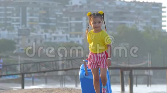 小可爱微笑的女孩正拿着一个手提箱在度假海滩上散步视频的预览图