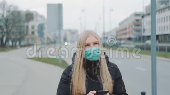 冠状病毒大流行戴着医用口罩的金发女人在智能手机上写信息视频的预览图