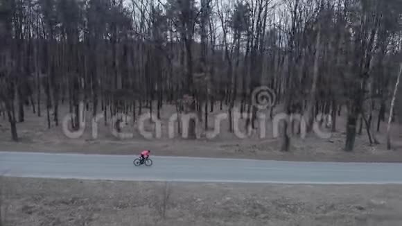 戴着头盔的女人骑着自行车在森林里的空车路上骑着自行车骑自行车的人在林地的隔离公路上骑得很快三重奏视频的预览图