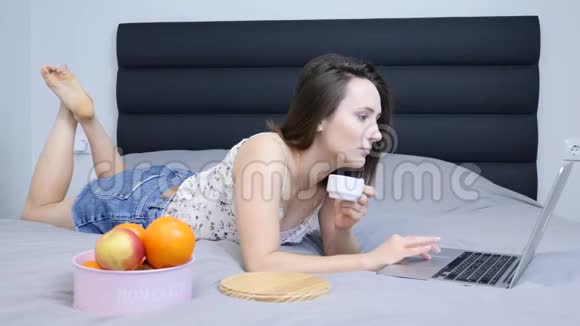 女人在酒店房间放松喝咖啡在笔记本电脑上在线阅读新闻穿着上衣和短裤的年轻迷人的千禧年视频的预览图