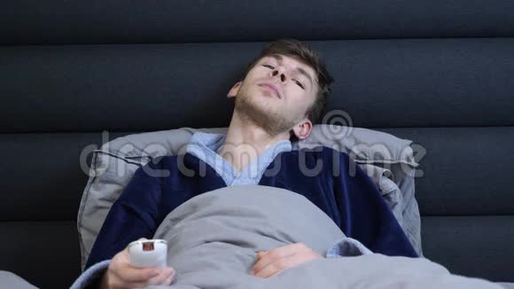 人躺在床上按遥控器上的按钮穿着浴袍的男人躺在床上看电视年轻人看电视新闻视频的预览图