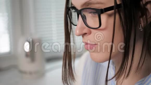 女孩吹鼻子年轻漂亮的戴眼镜的女人在厨房里看着笔记本电脑工作用纸打鼻子视频的预览图
