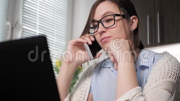 工作中的自由职业者漂亮的年轻女人戴着眼镜在厨房里打电话看着笔记本电脑同意视频的预览图