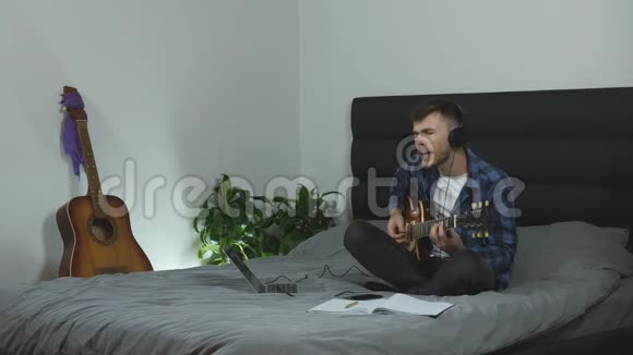 戴着耳机演奏吉他和唱歌的千禧一代穿着潘通蓝色t恤的年轻人在家里玩电吉他男性视频的预览图