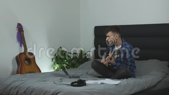 年轻的千禧一代弹吉他穿着Pantone蓝色t恤的魅力男人在现代livin坐在床上玩电吉他视频的预览图