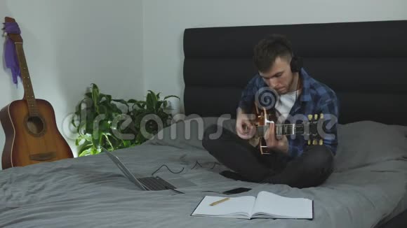 耳机里的吉他手用电吉他演奏音乐家在客厅的床上弹吉他年轻的白种人在潘视频的预览图