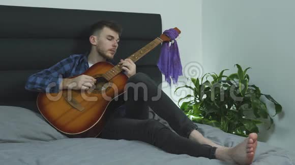 热的年轻音乐家在床上弹声吉他赤脚学习新歌音乐概念视频的预览图