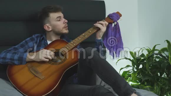 年轻英俊的胡子音乐家用吉他演奏情感摇滚流行歌曲坐在床上唱歌视频的预览图