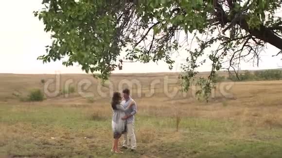 幸福的孕妇和她的丈夫在日落时在一棵大树附近的田野里视频的预览图