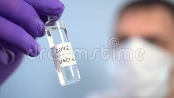 戴面膜的医生用蓝色背景的防护手套检查手中有冠状病毒疫苗的安瓿视频的预览图