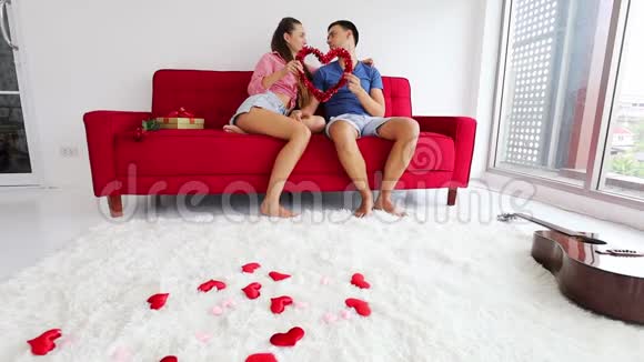 可爱的一对夫妇坐在红色的沙发上手里拿着红色的心环作为情人节爱情的象征视频的预览图