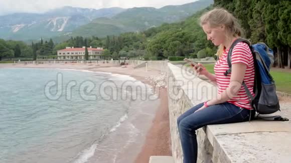 带袋包的Millenial女孩坐在海边带智能手机视频的预览图