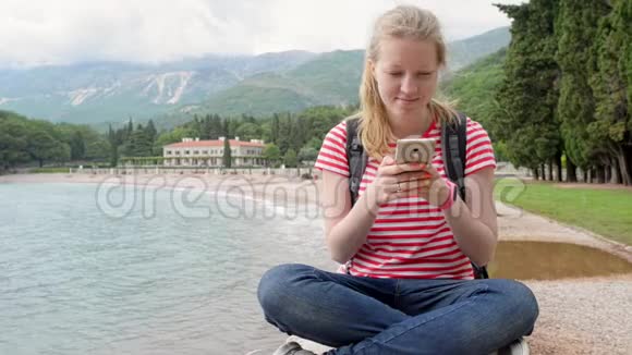 一个女孩和她的智能手机坐在海边的山脉和树木视频的预览图