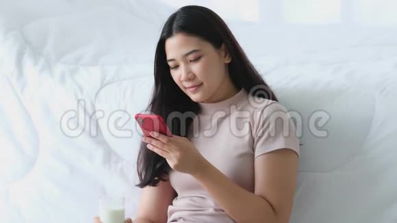 美丽的亚洲年轻女性坐在床边在卧室里用电话玩游戏视频的预览图