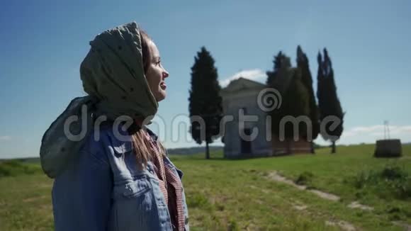 戴着头巾的漂亮女孩站在模糊的小教堂旁享受阳光视频的预览图