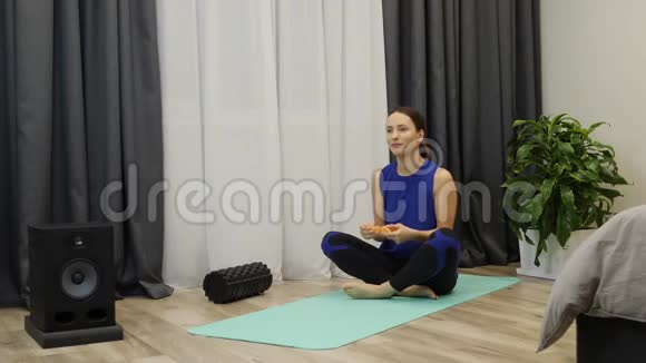 女人在家坐在瑜伽垫上吃水果莲花位置的年轻黑发女人吃柑橘穿着潘通蓝色瑜伽服的女性视频的预览图