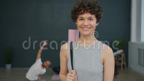 穿着运动服装手持瑜伽垫在工作室微笑的迷人女孩的肖像视频的预览图
