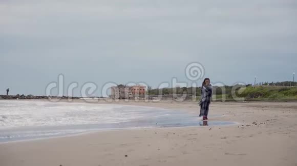 一个在阴天裹着毯子在海边散步的漂亮女孩视频的预览图