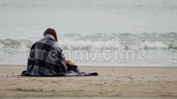 风和日丽的春日坐在海边的布鲁内特女孩裹着温暖的格子毯子大海上有波浪后视图视频的预览图