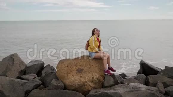 穿着黄色夹克的快乐的年轻女子坐在沙滩上的岩石上与海和天相对生活方式娱乐独处冥想视频的预览图