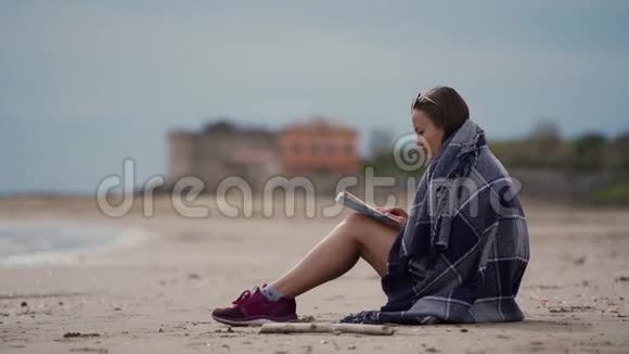 长头发的黑发女孩坐在海边穿着温暖的格子呢在阴天读着有趣的书侧视图视频的预览图