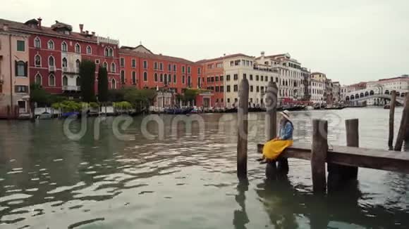 戴着帽子的女人穿着黄色裙子坐在威尼斯的木墩上意大利建筑视频的预览图