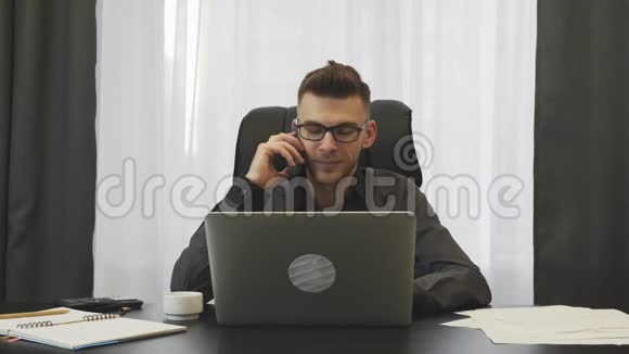 快乐商人在现代办公室打电话年轻兴奋的戴着眼镜的男人坐在办公桌前打电话M视频的预览图